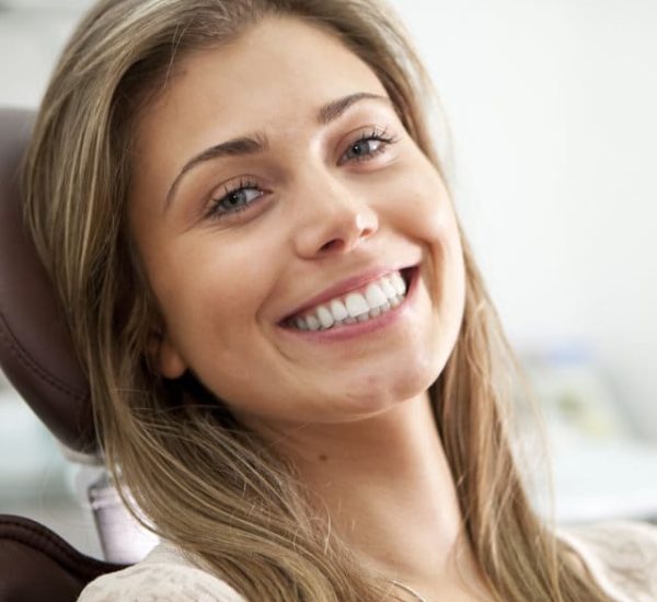 A bright toothy smile and eye contact from a pretty young woman proud to show off her perfect teeth while sitting in a dentist's chair.