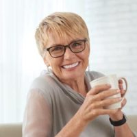 Portrait of happy mature woman in eyeglasses resting on sofa with cup of coffee and smiling at camera at home