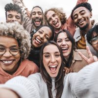 Multicultural community of young people smiling together at camera - Happy diverse friends taking selfie picture with smart mobile phone device - Friendship and human relationship concept